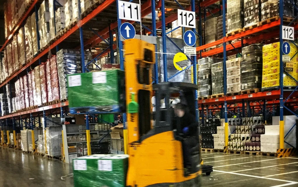 高い棚の並ぶ倉庫で、リーチフォークリフトが荷を運ぶ