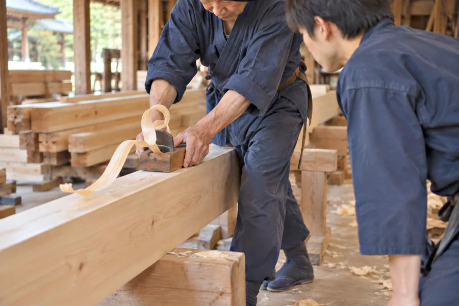 檜の梁にかんなをかける宮大工の手元を、弟子が肩越しに見て覚えようとしている