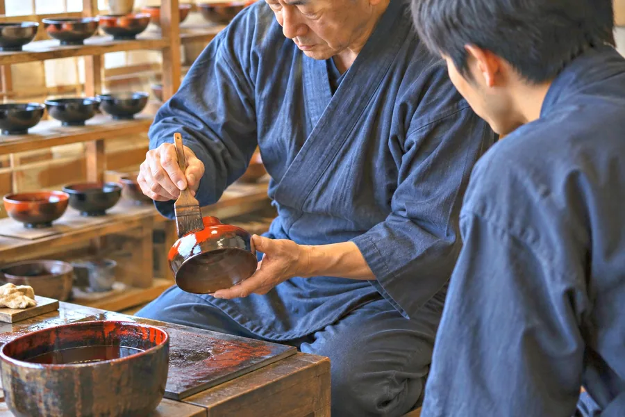 椀に漆を塗る職人の刷毛さばきを、弟子が肩越しに見て覚えようとしている