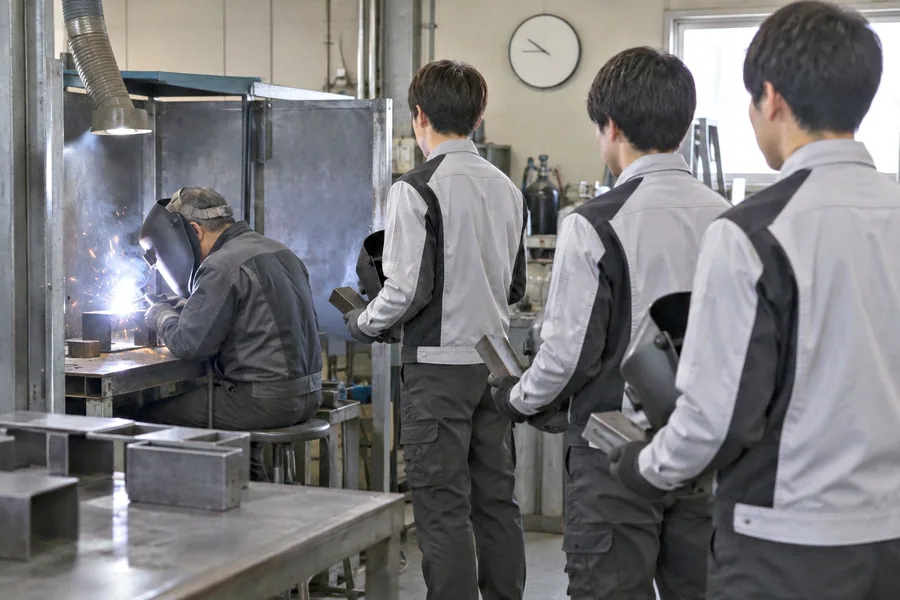 溶接する熟練者の手元を、新人が肩越しに見て覚えようとしている