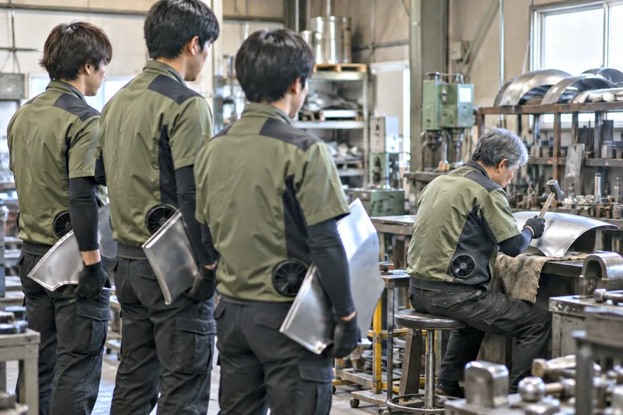 失敗した板金の板が並ぶ横で新人がたたき、熟練者は腕を組んで見ている